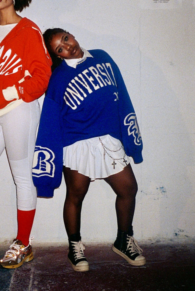 Two people wearing colorful sweatshirts and pants against a plain wall.